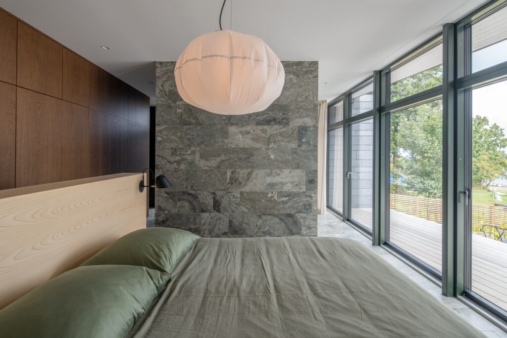 A large bed shown in a master bedroom with a marble separating wall, a large lampshade and glass doors leading to an outdoor patio.