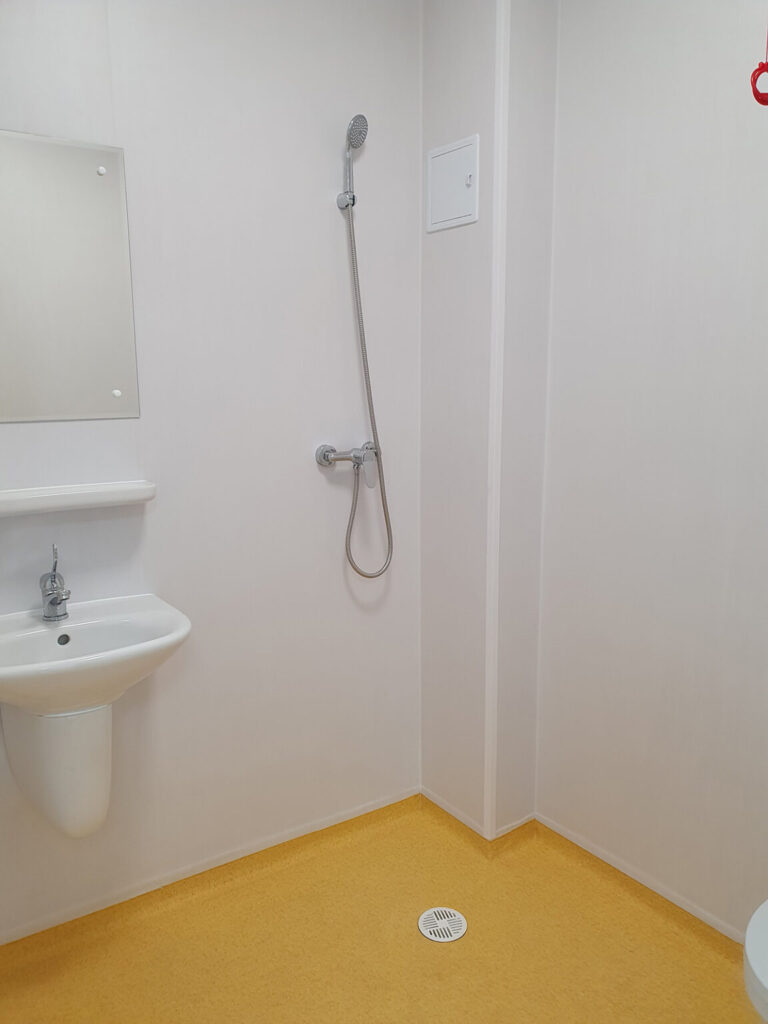A wetroom shower area with white walls and gold coloured vinyl flooring. There is a shower hose on the wall, and a sink and mirror to the left-hand-side. There is a small white drain in the floor.