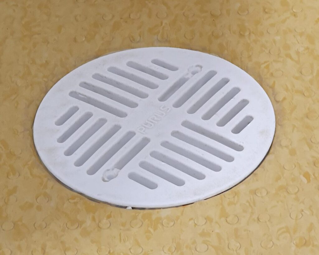Zoomed in image of a white plastic grate in a gold to beige vinyl floor.