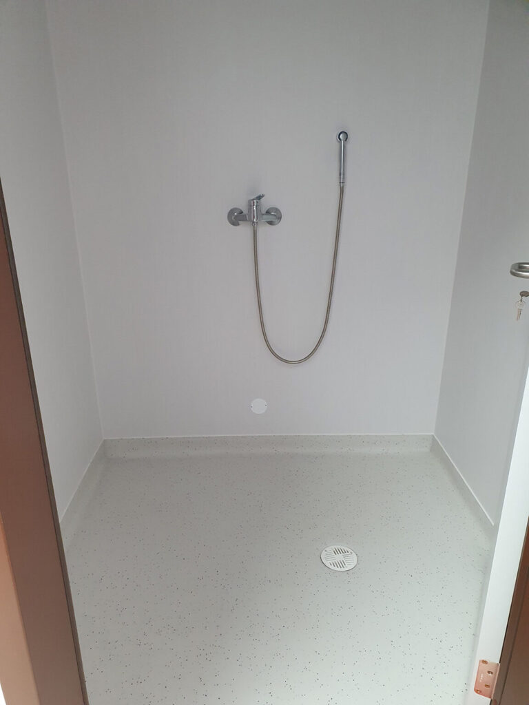 A shower area with a grey floor and white walls. A shower hose is on the wall, and a small white drain is in the floor.